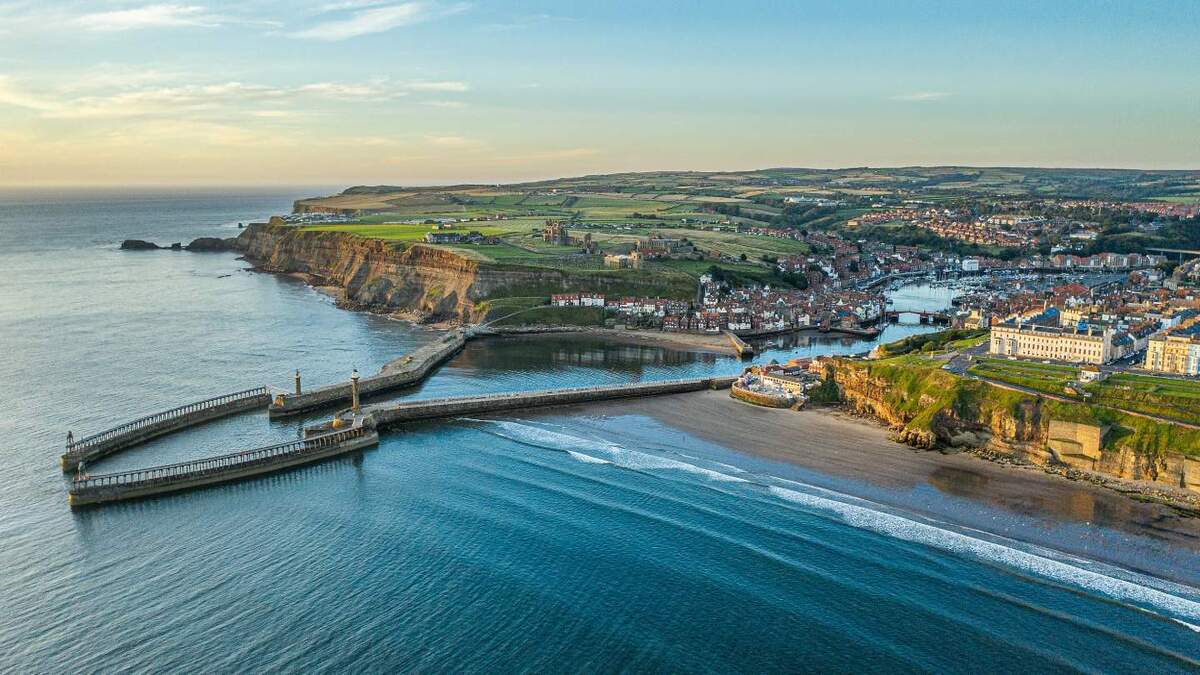 A stunning aerial view of Whitby, a coastal town in North Yorkshire, UK, featuring Whitby Abbey, the harbor, and the piers extending into the North Sea.