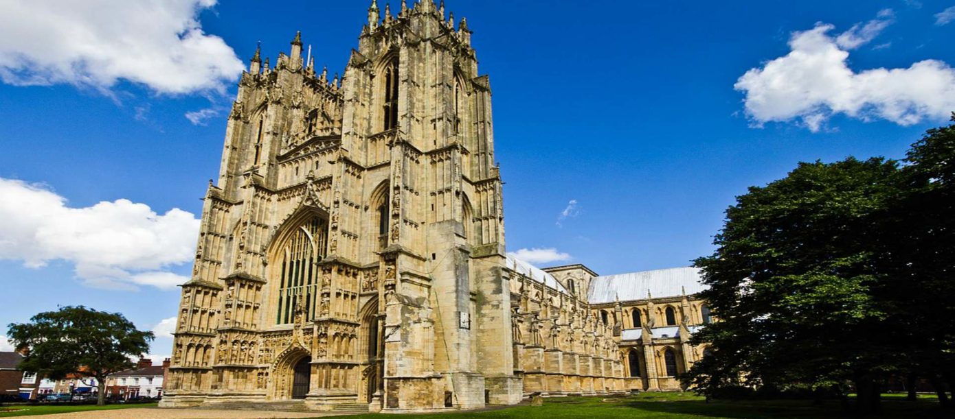 A stunning view of Beverley Minster, a grand Gothic-style church with intricate stonework and towering spires, set against a bright blue sky with scattered white clouds. Lush green trees and a well-kept lawn surround the historic structure.