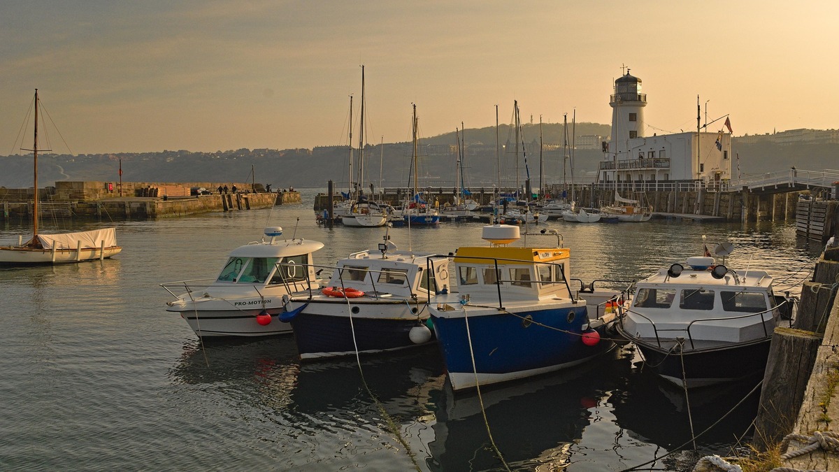 A scenic view of Scarborough Harbour in the evening, featuring boats docked in calm waters with a lighthouse in the background.