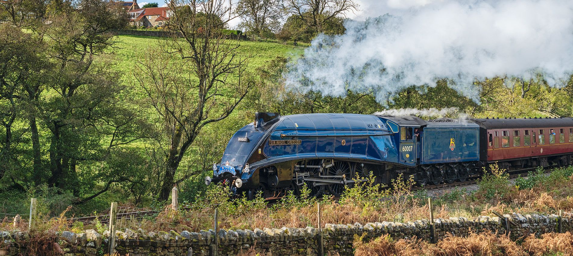 A striking blue steam locomotive, Sir Nigel Gresley, pulling vintage carriages through the scenic countryside of the North Yorkshire Moors Railway, with steam billowing into the sky.